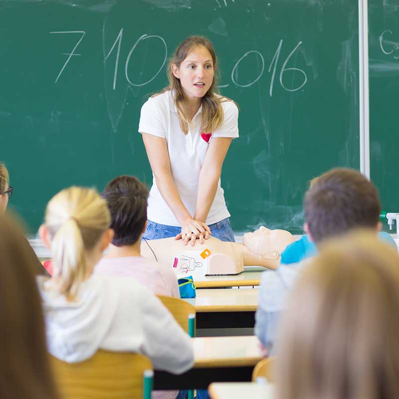 Person demonstrating CPR on a mannequin in a school classroom with a green chalkboard.
