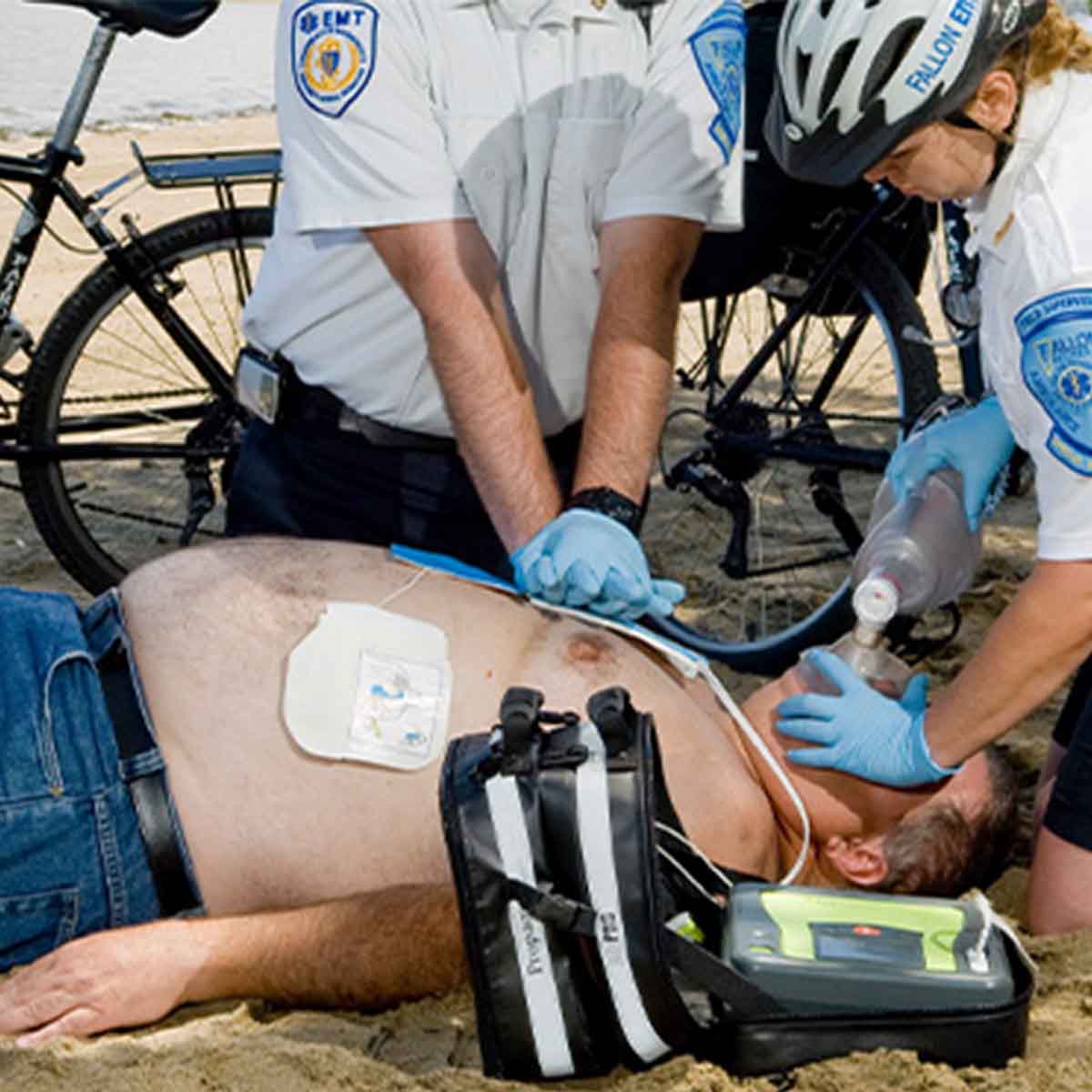 Person receiving CPR on a beach with emergency responders and medical equipment.