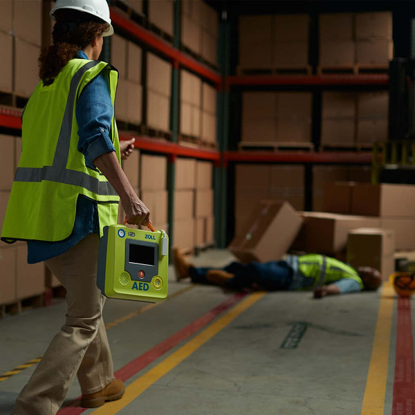Person in a warehouse setting with a defibrillator, another person lying on the ground, and boxes in the background.