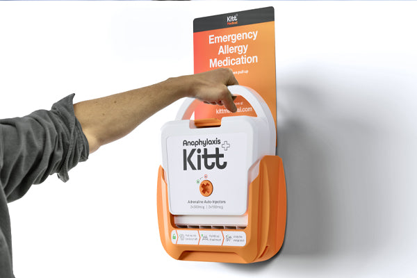 Person reaching to open a wall‑mounted Anaphylaxis Kitt emergency allergy medication box with adrenaline auto‑injectors in an orange holder