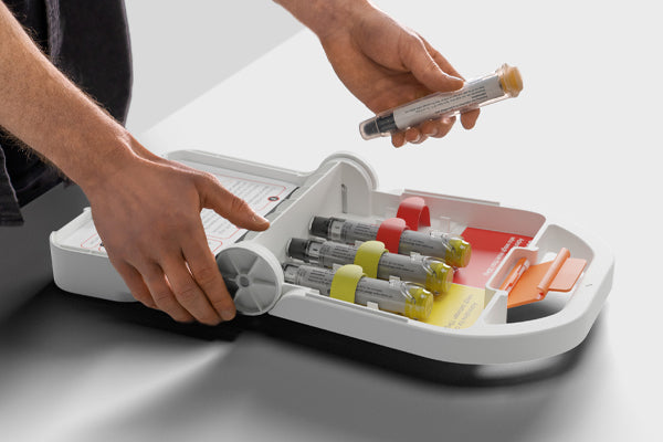 Hands opening an Anaphylaxis Kitt emergency allergy kit, showing multiple adrenaline auto‑injectors stored in colour‑coded compartments.
