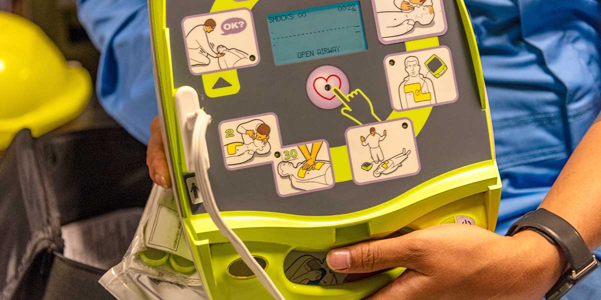 Automated External Defibrillator (AED) with user instructions displayed on a screen, held by a person wearing a hard hat.