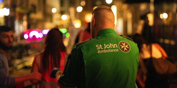 Person wearing a St John Ambulance green vest in an urban setting with blurred lights.
