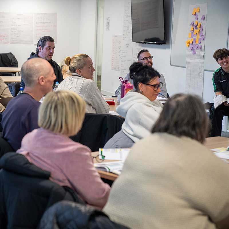 Group of people sitting around a table in a meeting room with a whiteboard and TV in the background.