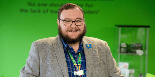 Man with glasses and a beard wearing a gray blazer and colorful shirt in front of a green screen with text.