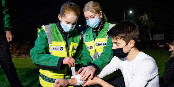 Two people in St. John Ambulance and Cadet uniforms assisting a person with a bandage at night.